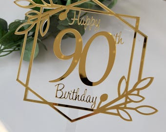Könnte beinhalten: Ein goldfarbener Kuchenaufsatz aus Acryl mit den Worten "Happy 90th Birthday" in eleganter Schrift. Das Design zeigt einen geometrischen Rahmen mit Blättern. Der Aufsatz hat einen transparenten Stiel.