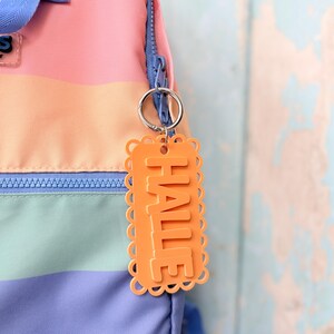 May include: An orange rectangular keychain with a scalloped edge and the name HALLE in raised block letters, attached by a silver metal ring to a pastel-colored striped backpack with a blue zipper and blue fabric handle.