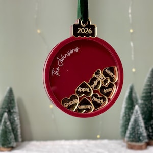 以下が含まれることがあります： 緑色のベルベットリボンが付いた赤い丸いクリスマスオーナメント。上部には2026年と刻印されています。前面には「The Johnsons」という文字があり、Granda、Mummy、Teddy、Jingles、Evie、Daddy、Jean、Grannyという名前が刻まれた小さな金色のハート型のチャームがいくつか入っています。
