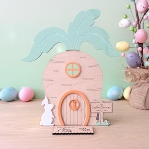Puede incluir: Casa de conejo de Pascua de madera con forma de zanahoria, con puerta y marcos de ventanas de color naranja claro. La casa tiene una parte superior de hojas verdes y un letrero que dice "The Easter Bunnys Magic Door". Un recorte de conejo blanco y una alfombra de bienvenida están al frente.