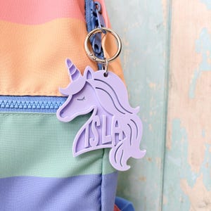 May include: A light purple unicorn-shaped keychain with the name ISLA embossed on the side, attached to a silver metal ring. The keychain is clipped to the blue zipper pull of a backpack featuring horizontal stripes in pastel pink, orange, and blue.