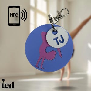 Könnte beinhalten: Ein runder Schlüsselanhänger in Blau und Lila mit einer Gymnastinnen-Silhouette und einem weißen Anhänger mit den Initialen "TJ" in Blau. Der Schlüsselanhänger hat einen silbernen Drehverschluss.