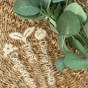 May include: Clear acrylic garden markers with white text. The markers are shaped like plants and feature the words "Coriander", "Mint", and "Cherry Tomato". Green leafy sprigs and a woven basket background complete the image.