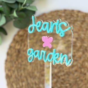 Può includere: Un paletto da giardino in acrilico trasparente con le parole "Jean's garden" in corsivo turchese. Una farfalla rosa è centrata tra le parole. Il paletto è progettato per l'uso all'aperto e aggiunge un tocco decorativo a qualsiasi giardino.