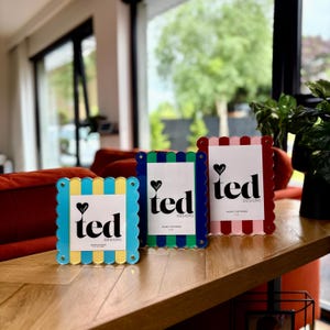 Peut inclure: Trois cadres photo colorés aux bords festonnés. Les cadres sont bleus, verts et rouges, chacun avec un insert blanc portant le texte "ted designs" et un cœur. Les cadres sont exposés sur une surface en bois.