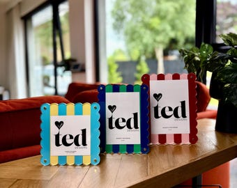 May include: Three colorful photo frames with scalloped edges. The frames are blue, green, and red, each with a white insert featuring the text "ted designs" and a heart. The frames are displayed on a wooden surface.