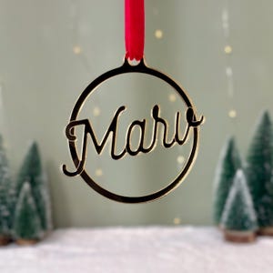 May include: A circular gold-toned ornament featuring the name Mary in a cursive script font. The ornament is suspended by a bright red ribbon loop at the top, creating a decorative piece with a reflective metallic finish and smooth edges.
