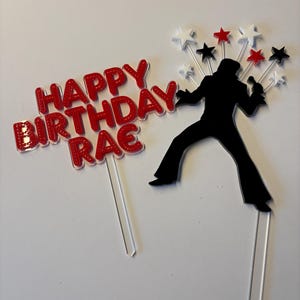 May include: A red and white birthday cake topper with the text "HAPPY BIRTHDAY RAE". A black silhouette of a singer with a microphone is also included, along with star accents.
