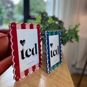 Peut inclure: Deux cadres photo décoratifs aux bords festonnés. L'un est rouge et rose, l'autre bleu et vert. Les deux cadres affichent le texte "ted DESIGNS" et un symbole de cœur. Les cadres sont magnétiques et mesurent 17.78 x 12.7 cm.