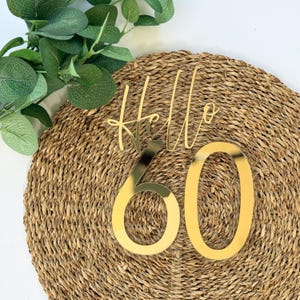 May include: A gold-colored cake topper with the word "Hello" in cursive script above the number "60". The topper is placed on a woven, circular mat with green foliage in the background. A celebratory decoration for a 60th birthday.