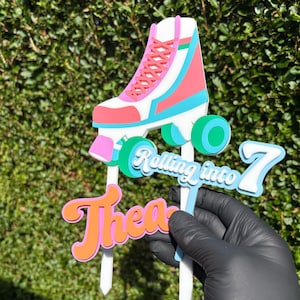 May include: A multi-layered cake topper featuring a white, pink, and teal roller skate, the text "Rolling into 7" in blue and white, and the name "Thea" in orange and pink, all mounted on white sticks for placement in a cake or dessert.