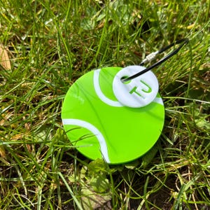 May include: A green tennis ball-shaped keychain with white stripes and a white tag. The tag has the initials "TJ" in green. The keychain is attached to a black and silver clasp, resting on green grass.