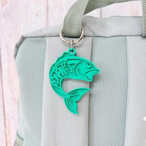 May include: A bright teal fish-shaped keyring featuring the name RONAN cut out in a bold, blocky font. The keyring is attached to a silver-toned metal split ring, which is secured to the light sage green fabric strap of a backpack.