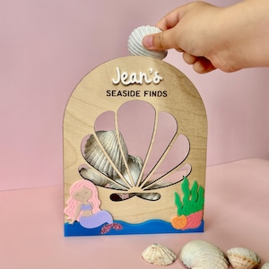 Può includere: Una scatola espositiva in legno a forma di arco con un design a conchiglia ritagliato e la scritta "Jean's SEASIDE FINDS". L'espositore è decorato con una sirena dai capelli rosa e alghe verdi, contenente diverse conchiglie nell'apertura.