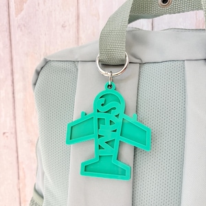 May include: A bright green acrylic aeroplane-shaped luggage tag featuring the name SAM cut out vertically along the fuselage. The tag is attached to a silver metal split ring, which is secured to the top handle of a light sage green backpack.