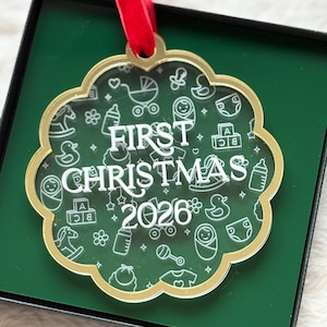 May include: A scalloped, clear acrylic ornament with a gold-toned border and a red velvet ribbon. The surface features white line art of baby items like bottles, strollers, and blocks, with the white text FIRST CHRISTMAS 2026 in the center.
