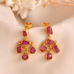 Peut inclure: Une paire de boucles d'oreilles pendantes dorées avec des pierres précieuses roses. Chaque boucle d'oreille présente une pierre rectangulaire en haut, avec un groupe de pierres précieuses roses en dessous.