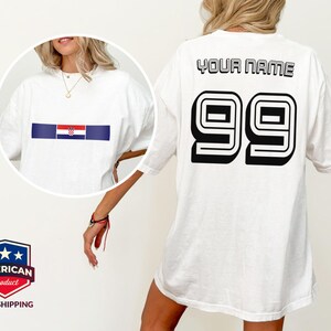 May include: A white t-shirt featuring a circular inset showing the front with a horizontal Croatian flag graphic. The back of the shirt displays the text YOUR NAME in black block letters above the large black outlined number 99 in a sports style.