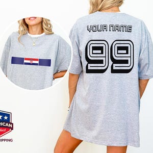 May include: A light gray t-shirt featuring a horizontal stripe with the Croatian flag on the front. The back displays the text YOUR NAME in black block letters above the large black number 99 with a white outline and a black drop shadow.