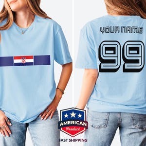 May include: A light blue t-shirt featuring the Croatian flag on the front chest. The back of the t-shirt displays the text YOUR NAME in black block letters above the large black number 99, which has a white outline and a 3D shadow effect.