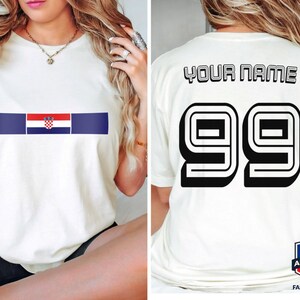 May include: A white short-sleeve t-shirt featuring a horizontal stripe with the Croatian flag on the front. The back of the shirt displays the text YOUR NAME in black block letters above the large black outlined number 99 in a sports style.