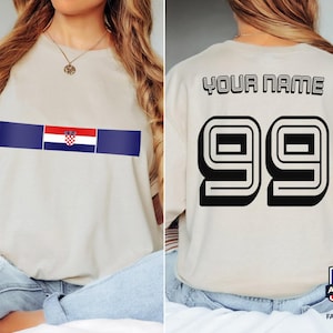 May include: A beige t-shirt featuring a horizontal stripe with the Croatian flag on the front. The back displays the text YOUR NAME in a black outlined font above the large black outlined number 99, styled like a sports jersey design.