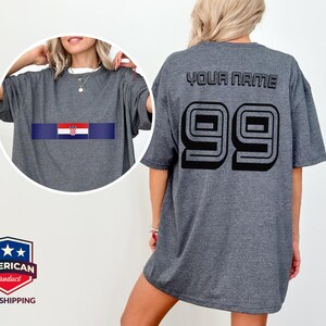 May include: A dark gray t-shirt featuring a circular inset showing the front with a horizontal Croatian flag graphic. The back of the shirt displays the text YOUR NAME in black block letters above the large black outlined number 99.