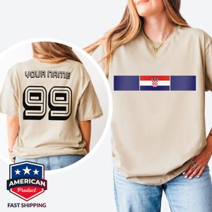 May include: A beige short-sleeved t-shirt featuring a horizontal stripe with the Croatian flag in the center. The back of the shirt displays the text YOUR NAME above the large black number 99 in a bold, block-style font with a white outline.