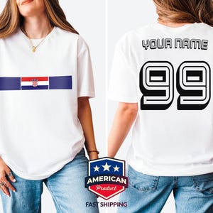 May include: A white short-sleeve t-shirt featuring a horizontal blue stripe with the Croatian flag in the center on the front. The back of the shirt displays the text YOUR NAME above the large black outlined numbers 99 in a bold athletic font.