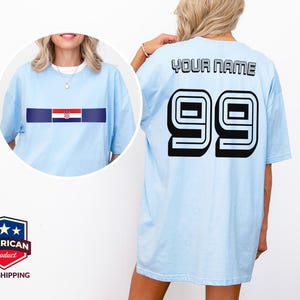 May include: A light blue t-shirt featuring a horizontal stripe with the Croatian flag on the front and the text YOUR NAME above the number 99 in large black block lettering on the back. The shirt has short sleeves and a crew neckline.