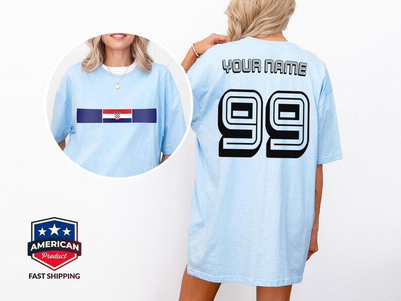 May include: A light blue t-shirt featuring a horizontal stripe with the Croatian flag on the front and the text YOUR NAME above the number 99 in large black block lettering on the back. The shirt has short sleeves and a crew neckline.
