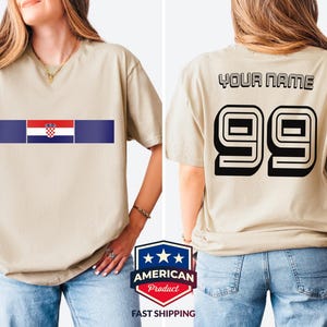 May include: A beige short-sleeve t-shirt featuring a horizontal graphic on the front with the Croatian flag centered between two dark blue bars. The back of the shirt displays the text "YOUR NAME" above the large black outlined number "99".
