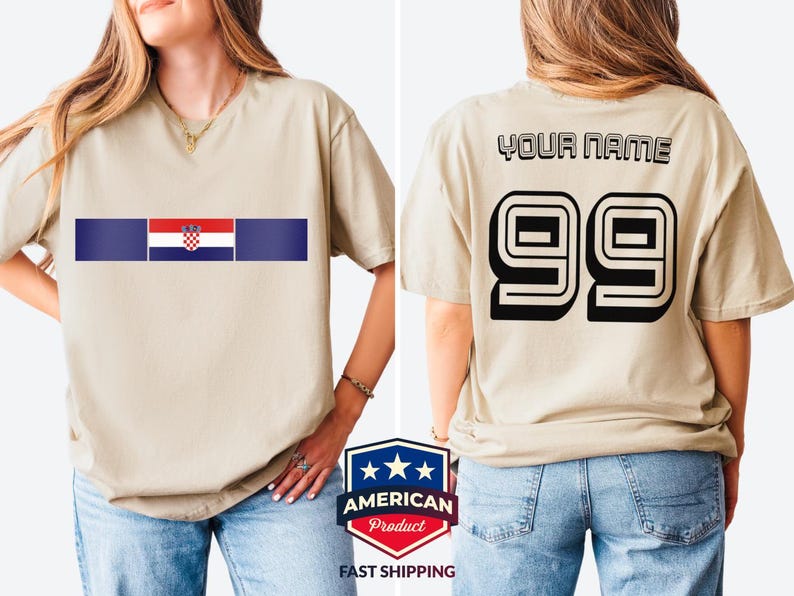 May include: A beige short-sleeve t-shirt featuring a horizontal graphic on the front with the Croatian flag centered between two dark blue bars. The back of the shirt displays the text "YOUR NAME" above the large black outlined number "99".