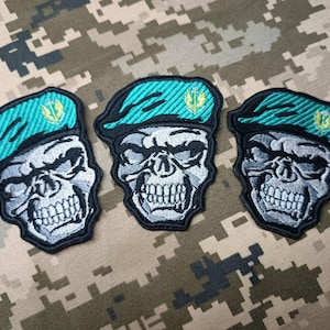 Puede incluir: Tres parches bordados con una calavera que lleva un gorro verde. Cada parche tiene un símbolo amarillo en el gorro.