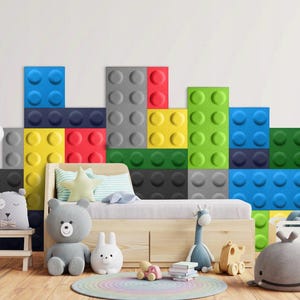 Könnte beinhalten: Ein Kinderzimmer mit einem Bett und einer Wand, die mit bunten, 3D-Lego-ähnlichen Blöcken verziert ist. Die Blöcke sind in Blautönen, Grün, Gelb, Rot und Grau gehalten. Auf dem Boden liegen Stofftiere und neben dem Bett steht eine Spielzeuggiraffe.