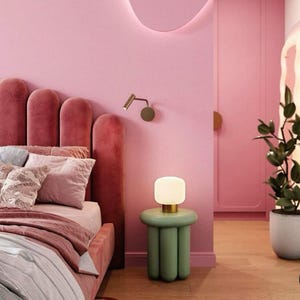Può includere: Un tavolino cilindrico verde salvia con base scanalata e piano arrotondato, sormontato da una piccola lampada in vetro a coste con base dorata. Il tavolino è posizionato accanto a un letto in velluto rosa con testiera smerlata.