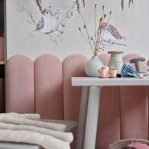 May include: A pastel-toned children's room featuring a pink upholstered wall and a white table. The table displays a vase, miniature teacups, and a paper fan. A plush octopus toy and a woven basket are also present.