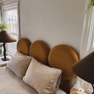 Puede incluir: Una cama con una cabecera hecha de tres grandes cojines redondos de terciopelo amarillo mostaza. La cama está hecha con ropa de cama de lino blanco y dos almohadas.