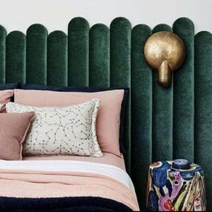 Può includere: Una testiera in velluto verde scuro con pannelli verticali arrotondati, dotata di una lampada da parete circolare in ottone. Il letto è decorato con cuscini rosa cipria, un cuscino bianco con motivo a costellazioni blu e una coperta rosa.