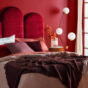Può includere: Una camera da letto con una testiera in velluto rosso intenso composta da due pannelli ad arco. Il letto è coperto da biancheria a strati, tra cui una coperta testurizzata marrone scuro, un piumino color cuoio e cuscini in tonalità rosso.