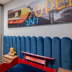 Può includere: Una testiera in velluto blu a pannelli verticali posizionata dietro una struttura letto rossa. Sopra la testiera è appesa una stampa su tela rettangolare con un'auto gialla, un'insegna al neon arancione OPEN e il testo 24/7 e On Route 66.