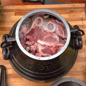 Peut inclure: Un kazan en fonte noir, un pot de cuisson traditionnel, rempli de viande crue et d'oignons tranchés. Le kazan est posé sur une surface en bois, avec un couteau et des herbes visibles en arrière-plan. Le pot a des poignées et des motifs décoratifs.