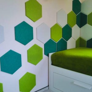 Op de afbeelding: Een witte muur met een hoofdeinde van een bed dat is bekleed met groene stof. De muur is versierd met zeshoekige akoestische panelen in tinten groen en wit.