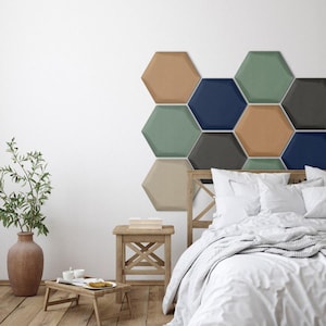 May include: A collection of hexagonal wall panels in shades of tan, sage green, navy blue, and charcoal gray arranged in a honeycomb pattern above a wooden bed frame with white bedding, a wooden side table, and a terracotta vase.