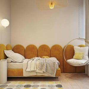 Può includere: Un letto imbottito color giallo senape con testiera smerlata, biancheria da letto bianca e una coperta in maglia grossa. Una sedia a bolla in acrilico trasparente pende dal soffitto con cuscini bianchi e gialli, accanto a un tavolino cilindrico bianco.