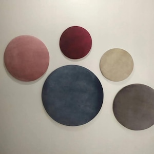 Puede incluir: Cinco piezas redondas de decoración de pared en varios colores y texturas. Las piezas están dispuestas en forma de semicírculo en una pared blanca. Los colores son rosa, rojo, beige, azul y gris.