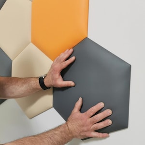 May include: A close-up of a wall covered in hexagonal acoustic panels. The panels are in shades of beige, orange, and gray. A person's hands are shown pressing on the gray panel.