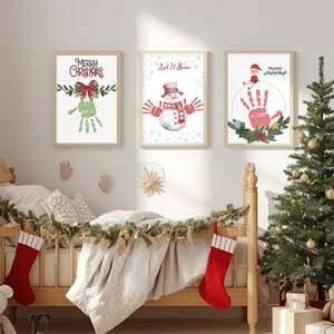 Könnte beinhalten: Drei gerahmte Weihnachtskunst-Drucke mit festlichen Designs. Einer zeigt "Merry Christmas" mit einem Handabdruck, ein anderer "Let It Snow" mit einem Schneemann, und der dritte "Merry Christmas" mit einem Handabdruck. Ein Weihnachtsbaum und ein Kinderbett sind im Vordergrund.