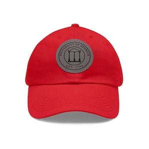 May include: A bright red baseball cap featuring a circular patch with the words "UNIVERSITY OF GEORGIA" and "EST. 1785" around a graphic of a building. The cap is made of a soft material and has a curved brim.
