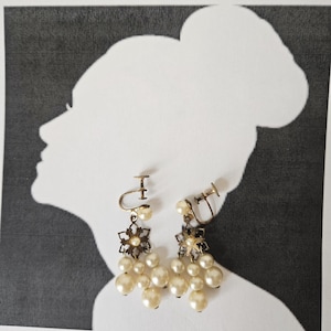 May include: Vintage clip-on earrings with a floral design and dangling faux pearls. The earrings have a gold-tone metal finish and are set against a black and white silhouette background. The earrings are a classic accessory.